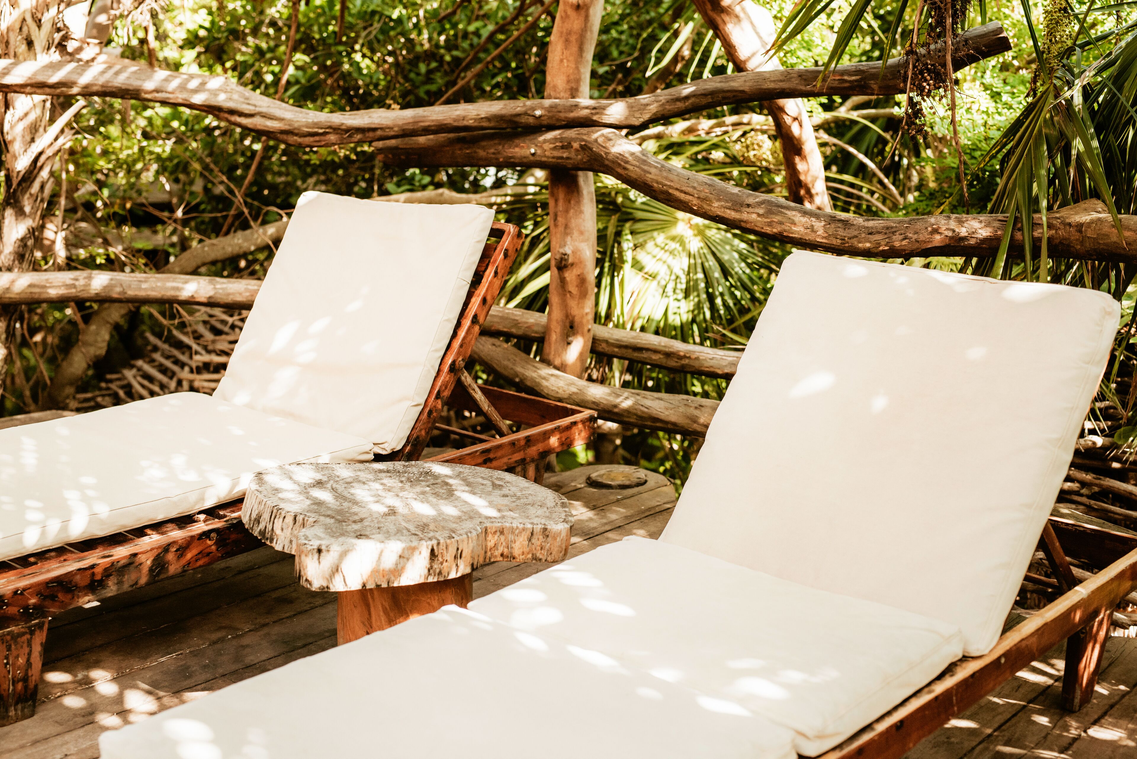 Two wooden lounge chairs with white cushions, positioned on a wooden deck surrounded by natural foliage.