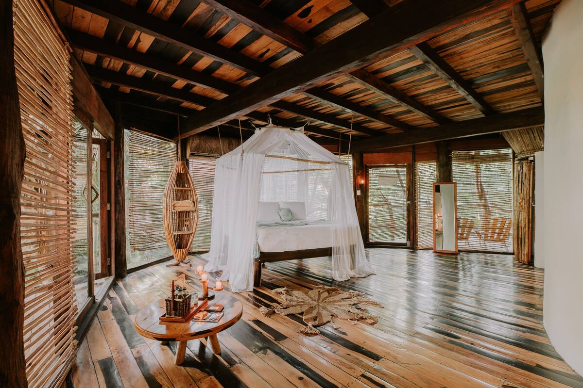 A room with a bed under a canopy, wooden floors, and large windows with blinds.