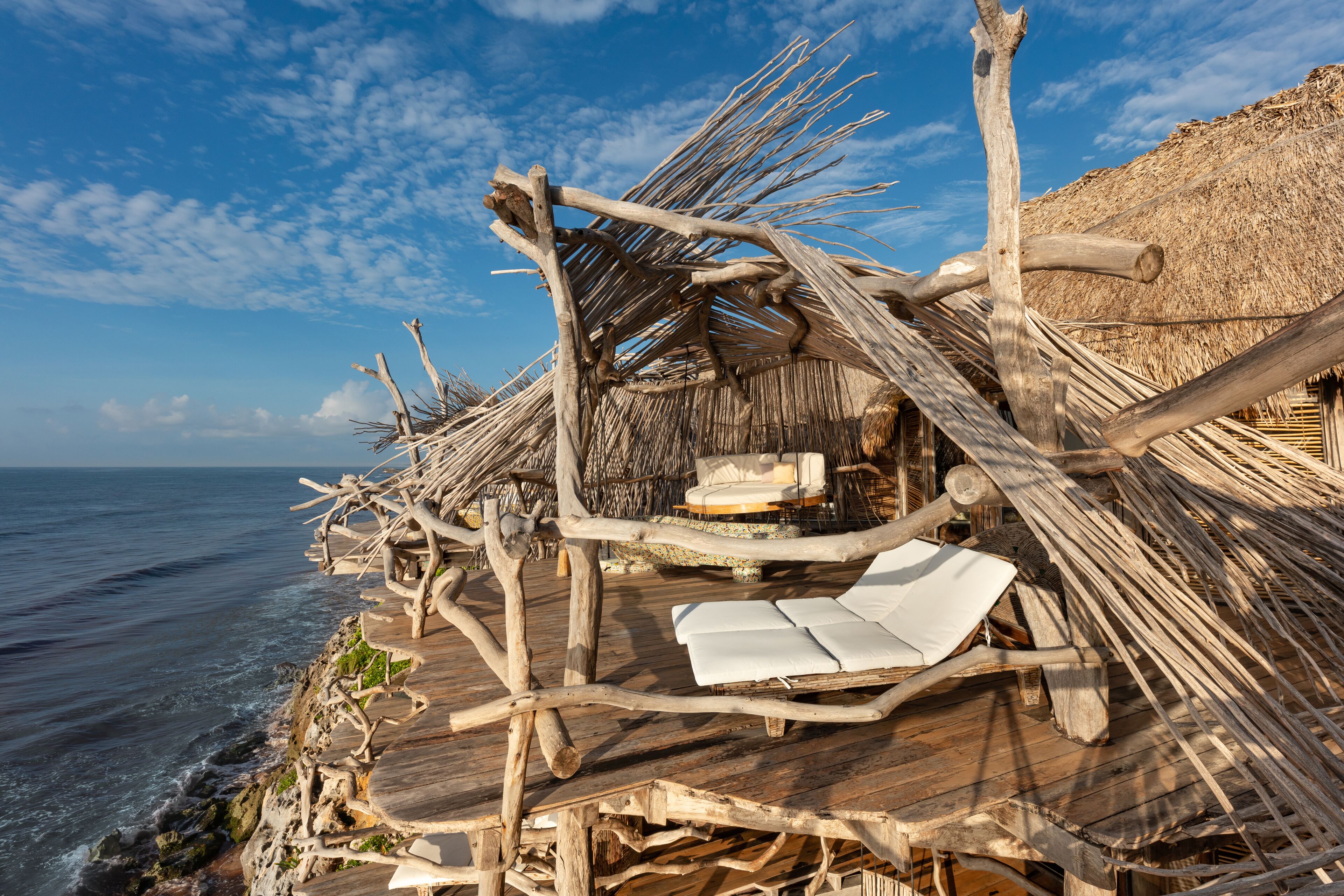 Una terraza de madera con techo de paja, una silla de lounge blanca y vista al océano.