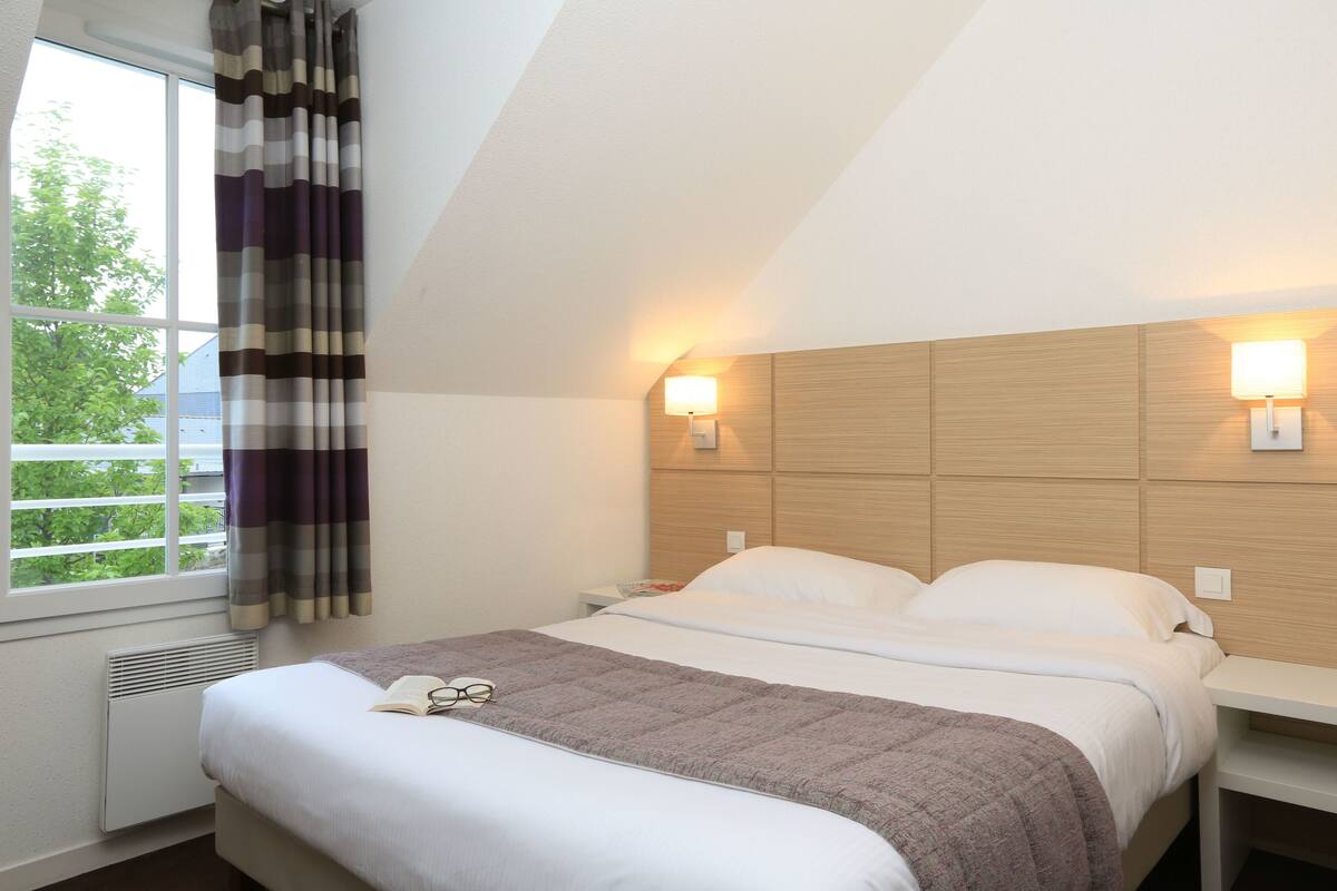 A hotel room with a bed, bedside tables, a window with curtains, and wall-mounted lights.