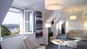 A modern hotel room with a large window offering a view of a river and bridge, a sofa, a coffee table, and a lamp. - Domaine de Pont Aven Art Gallery Resort (Pont-Aven)