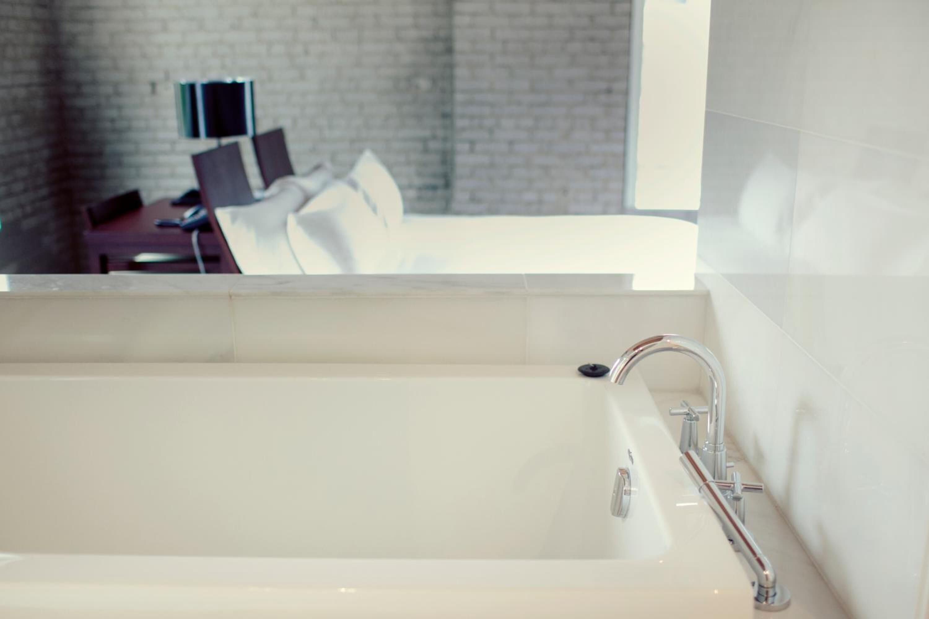 A modern bathroom with a large bathtub, a mirror, and a view of a room with a bed and a desk.