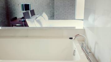 A modern bathroom with a large bathtub, a mirror, and a view of a room with a bed and a desk.