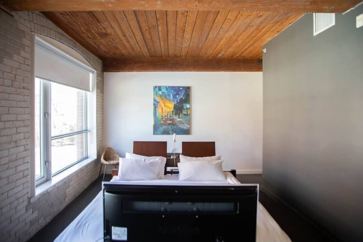 Une chambre avec un plafond en bois, une grande fenêtre, un lit avec du linge de lit blanc, un tableau au mur et une chaise.