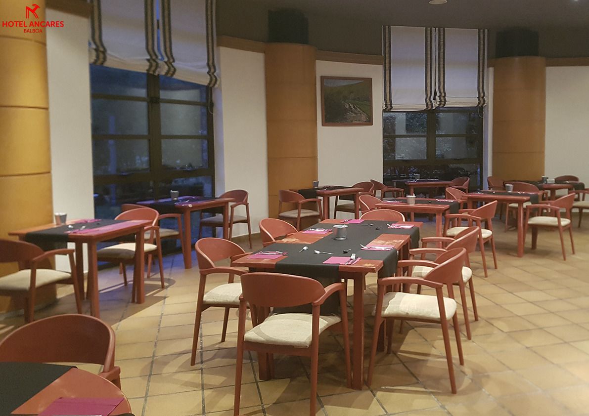 Un comedor de hotel con varias mesas y sillas, ventanas amplias y un cuadro en la pared.