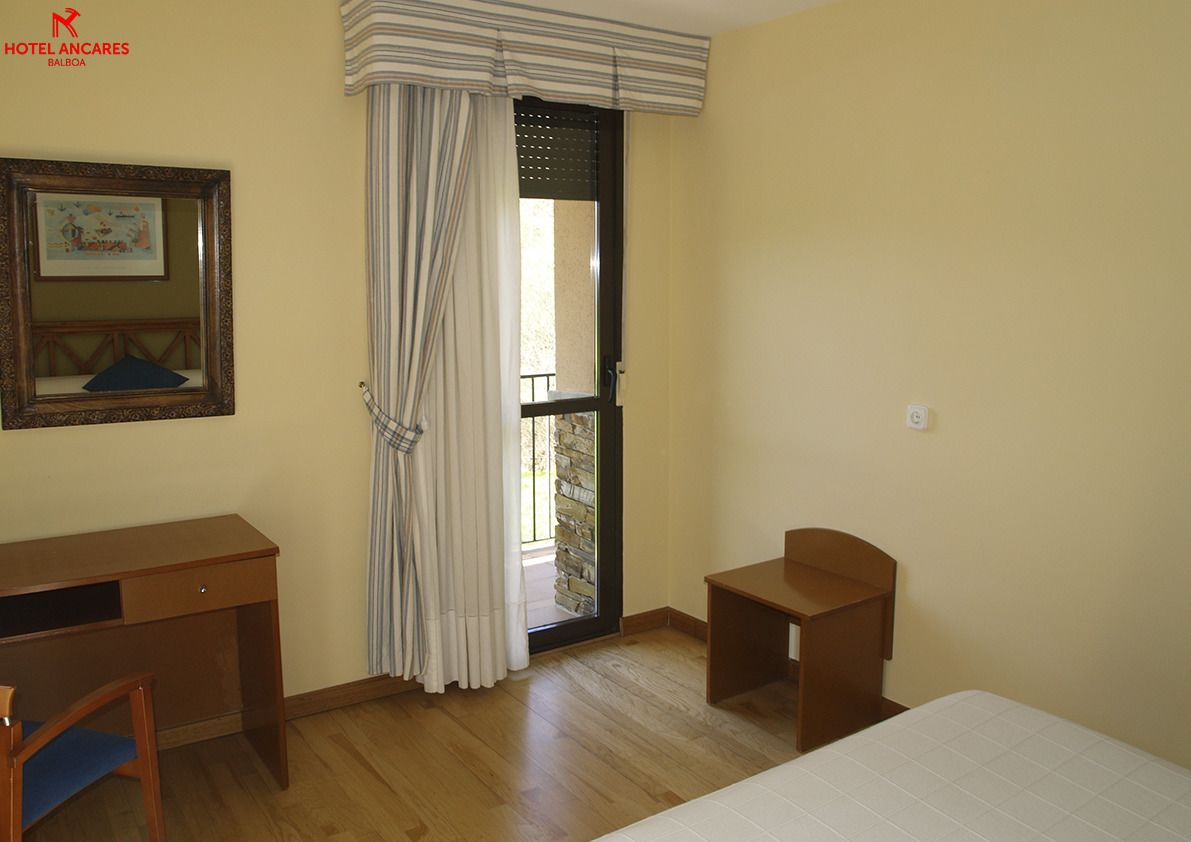 A hotel room with a wooden desk, a small wooden table, a bed, a window with curtains, and a framed picture on the wall.
