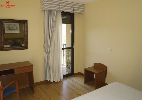Una habitación de hotel con un escritorio de madera, una mesita pequeña de madera, una cama, una ventana con cortinas y un cuadro en la pared.