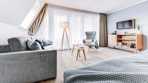 A modern hotel room with a grey sofa, a small round table, a floor lamp, a grey armchair, a bookshelf, and a TV mounted on the wall. - Hotel Muschelgrund (Cuxhaven)