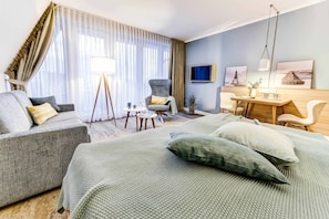 A hotel room with a bed, a sofa, a desk, and a chair. There is a lamp, a small table, and a plant.