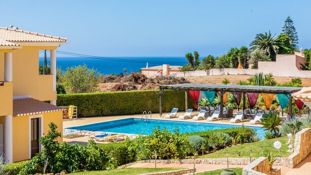 Un área de piscina con sillones de descanso, una pérgola con banderas de colores y vista al mar.