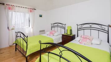 Quarto com duas camas, cada uma com colcha verde e almofadas brancas. Há uma janela com cortinas brancas e um pequeno quadro na parede.