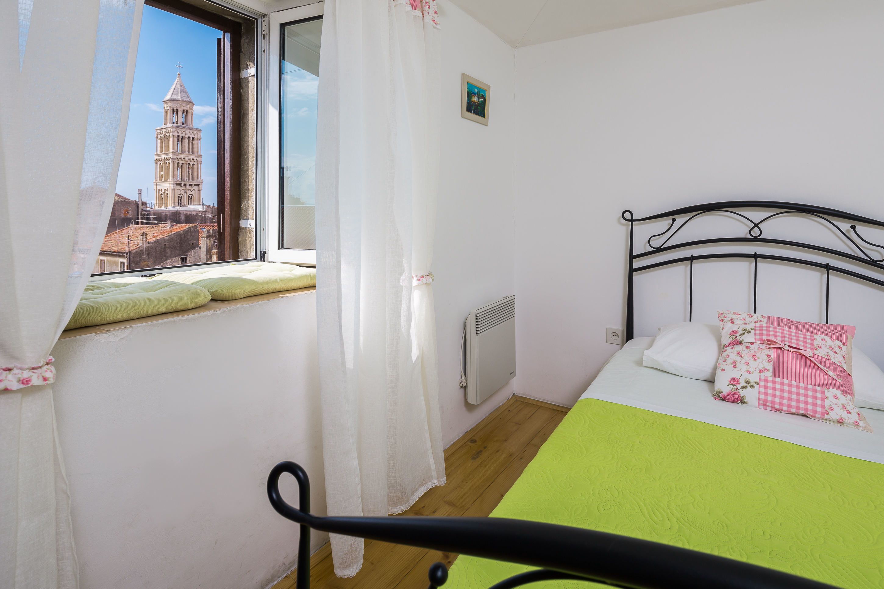 Un dormitorio con cama, ventana con vista a una torre, un radiador y un cuadro pequeño en la pared.