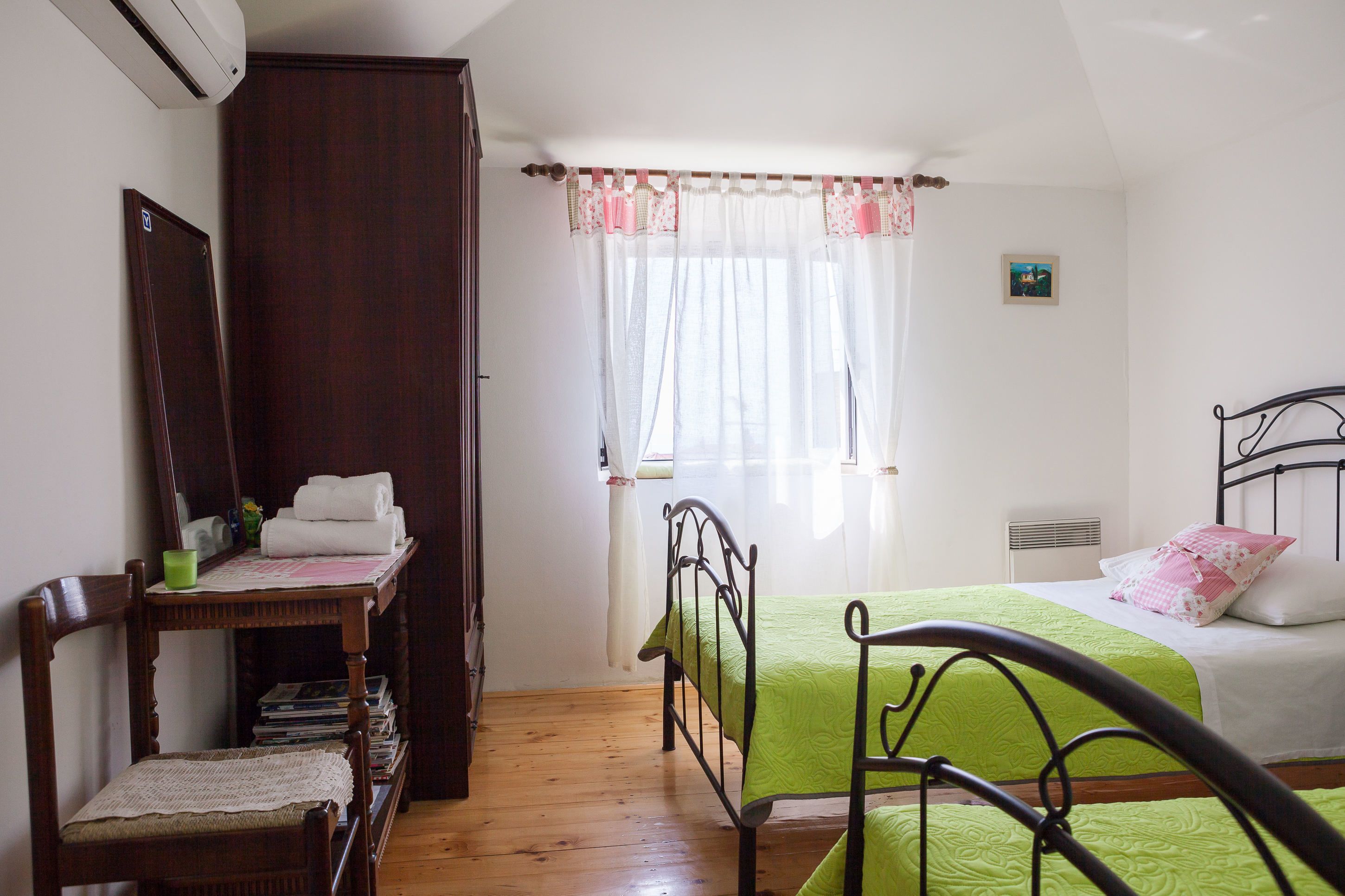 Ein Schlafzimmer mit einem Bett, einem Sessel, einem kleinen Tisch mit Spiegel, einem Kleiderschrank und einem Fenster mit Vorhängen.