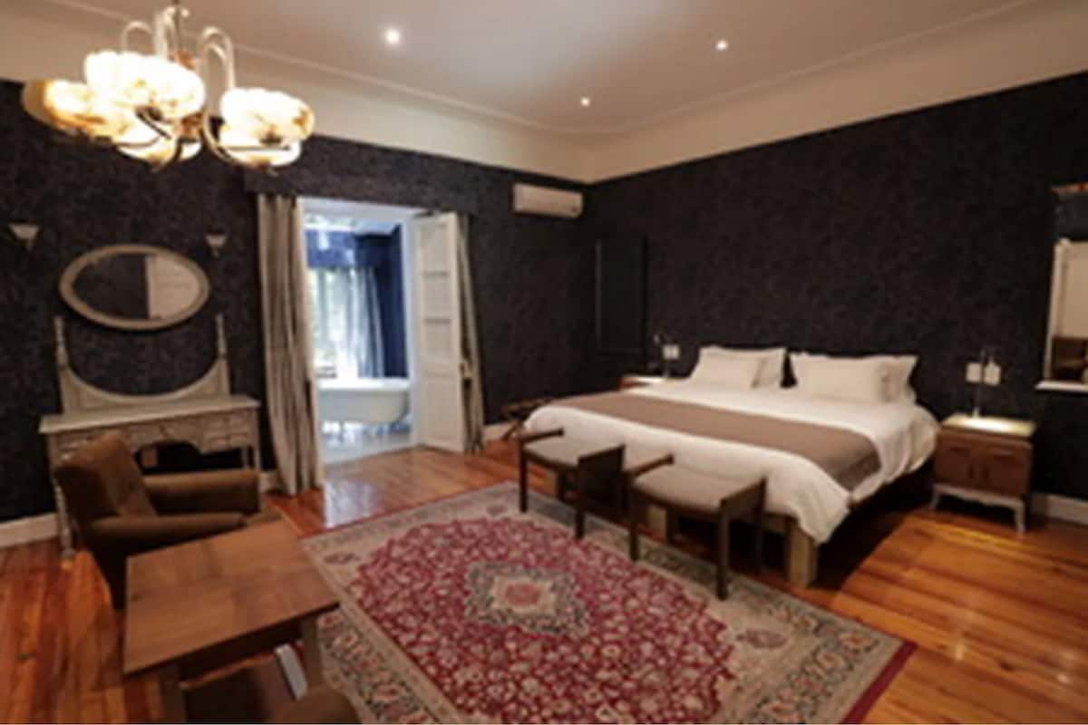 Ein Schlafzimmer mit einem großen Bett, Holzboden, einer Chandelüster, einem Spiegel und einer Tür zu einem anderen Raum.