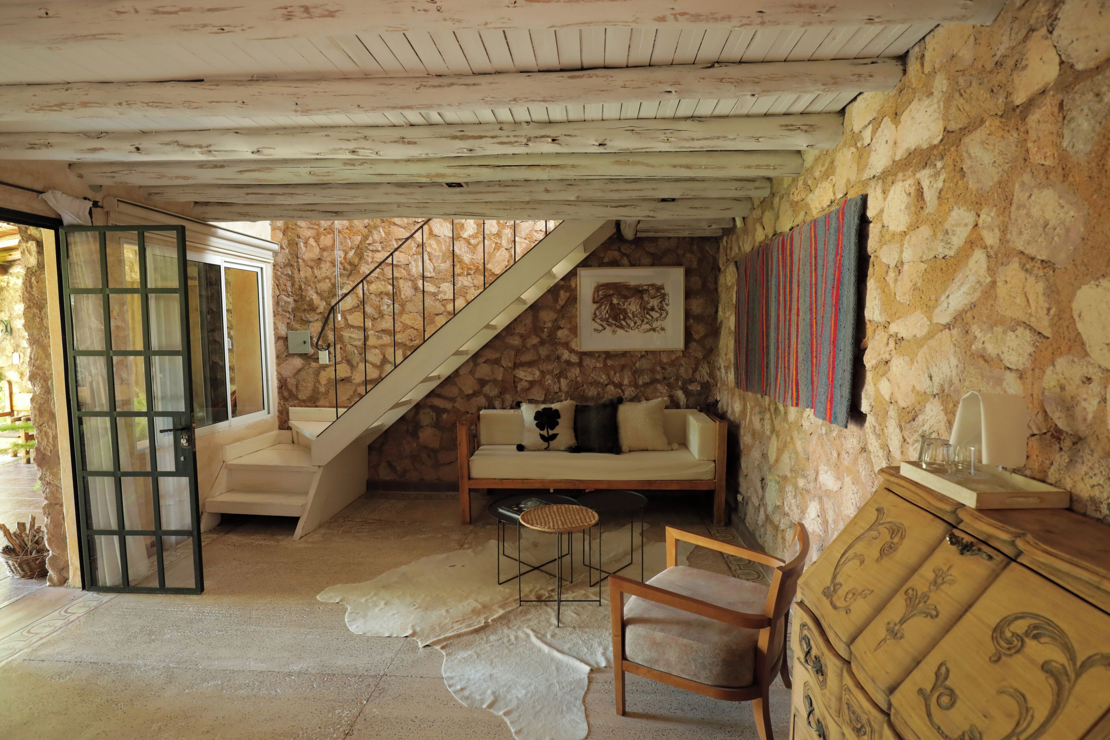 A rustic room with a stone wall, a wooden staircase, a sofa, a chair, and a small table.