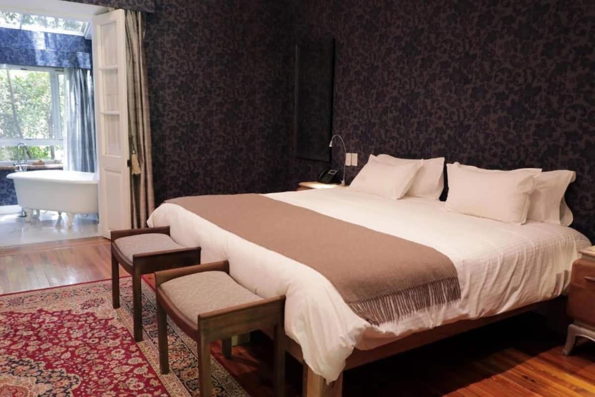 Ein Schlafzimmer mit einem großen Bett, einer Sitzecke mit einem Sessel, einem Nachttisch, einem Wandtelefon und Blick auf eine Badewanne und Grünflächen.