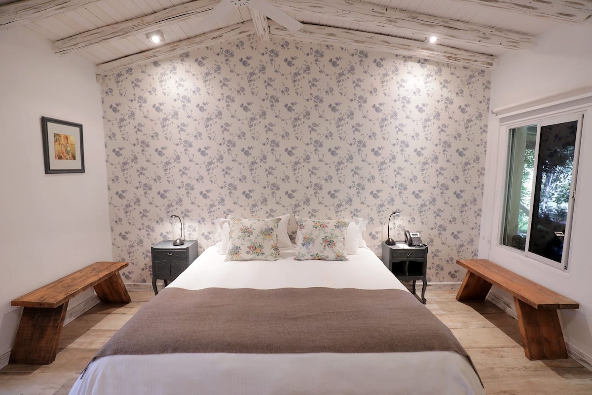 Ein Schlafzimmer mit einem großen Bett, floralem Wandpapier, zwei Nachttischen mit Lampen und einem Fenster.