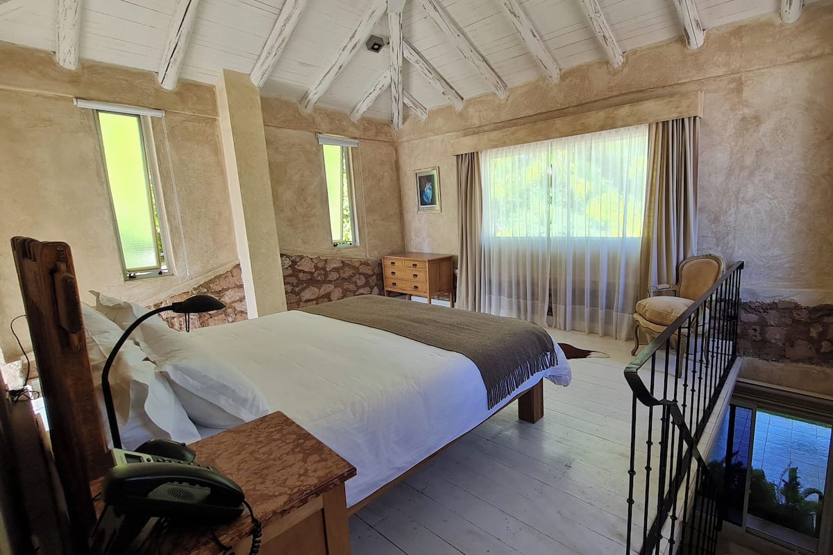 Ein Schlafzimmer mit einem großen Bett, einem Nachttisch aus Holz mit Telefon, einer Steinwand, einem Fenster mit Vorhängen und einem Stuhl am Fenster.