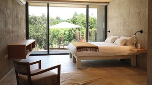 A bedroom with a bed, a desk, and a chair. There is a large window with a view of trees and a white umbrella. -  Finca Adalgisa Wine Hotel (Chacras de Coria)