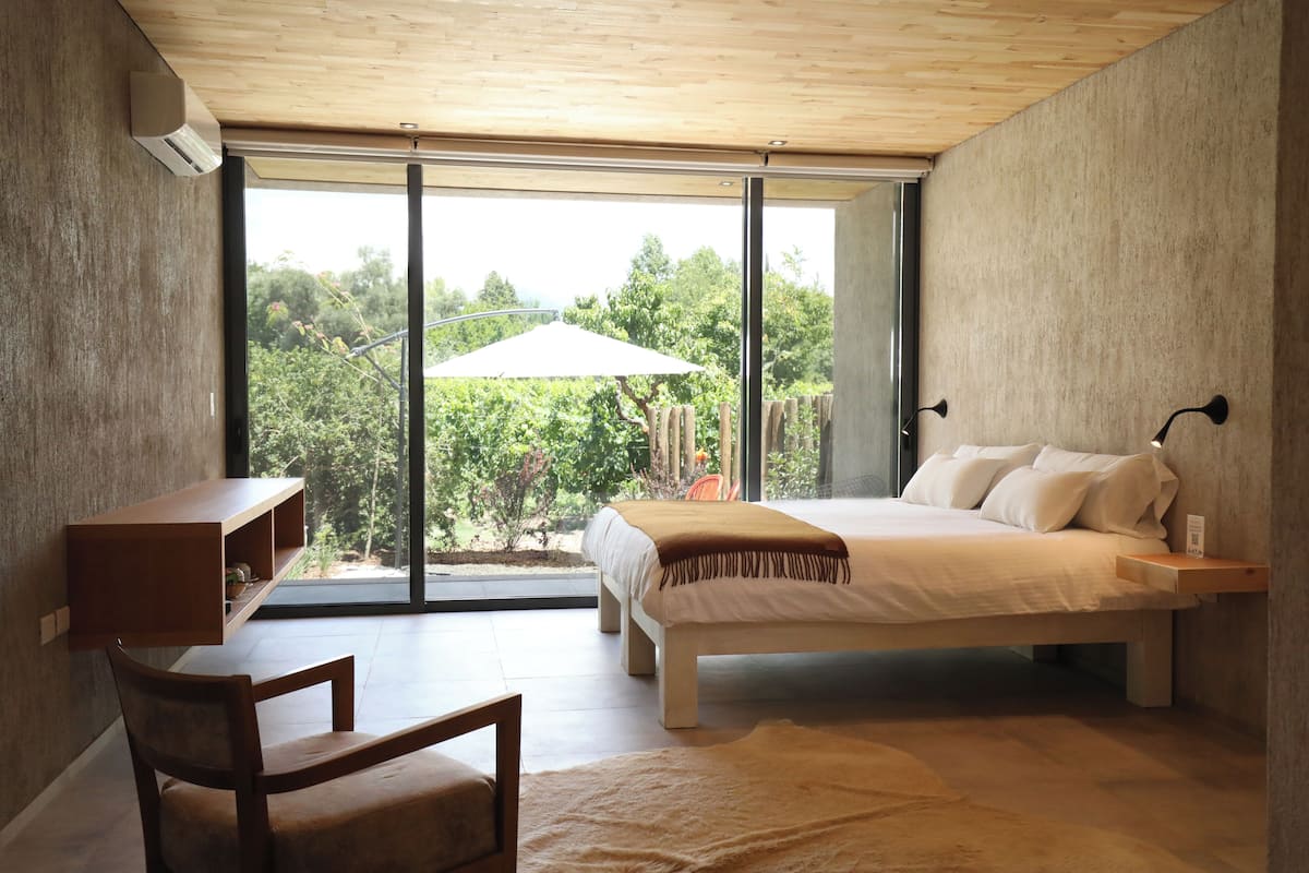Ein Schlafzimmer mit einem Bett, einem Schreibtisch und einem Stuhl. Ein großes Fenster bietet Blick auf Bäume, und ein weißer Sonnenschirm ist zu sehen.