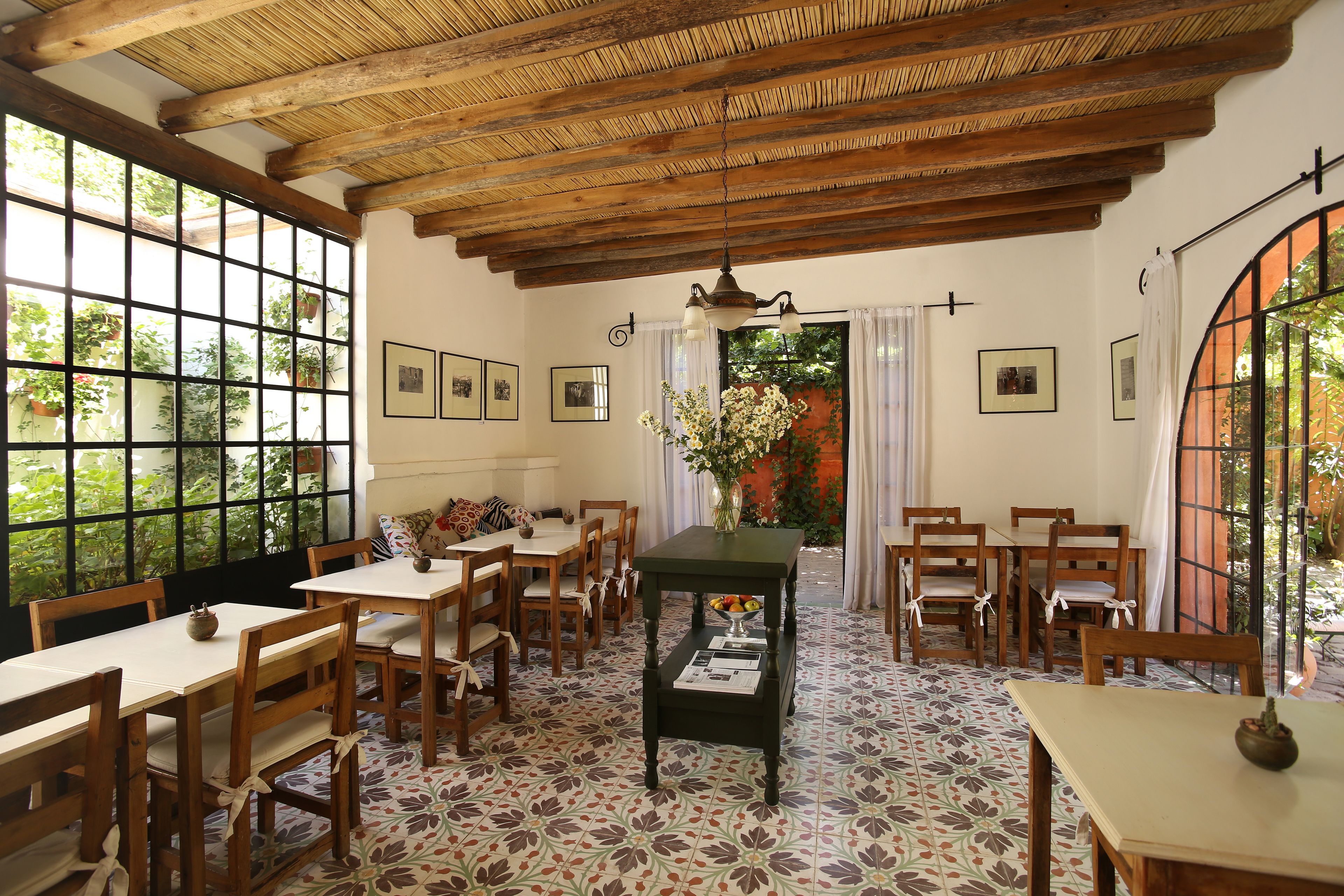Un comedor con mesas y sillas de madera, una mesa central con un jarrón de flores y ventanas amplias con vistas al follaje.