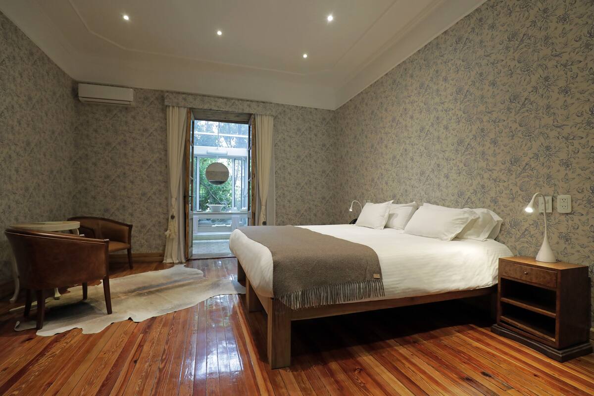 Ein Schlafzimmer mit Holzboden, einem Bett mit weißer Bettwäsche, einem Nachttisch mit Lampe, einem Sessel und einem Fenster mit Vorhängen.