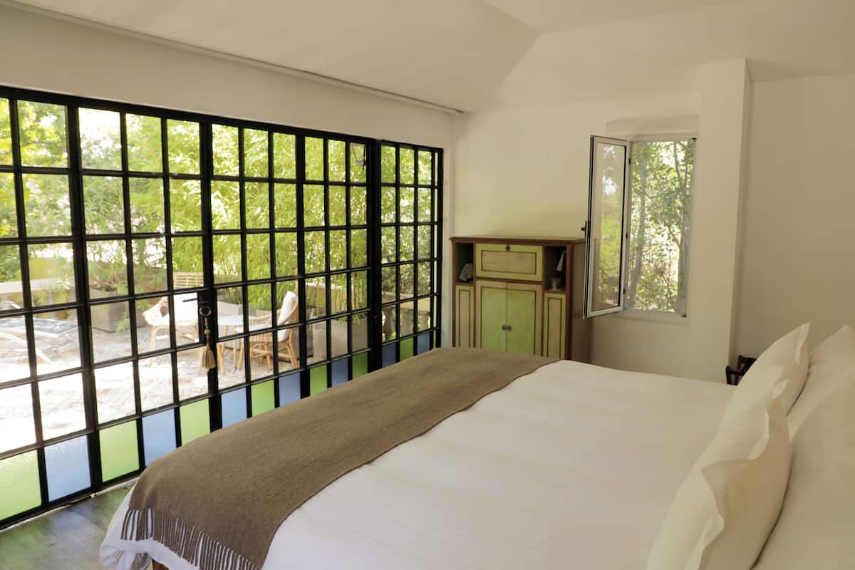 Ein Schlafzimmer mit großem Fenster, einem Bett, einem Nachttisch und Blick ins Freie.
