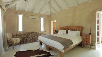 Un dormitorio con una cama grande, cabecera de madera, pared de piedra, alfombra de cuero de vaca, una silla y una mesita de noche con lámpara.