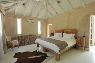 Un dormitorio con una cama grande, cabecera de madera, pared de piedra, alfombra de cuero de vaca, una silla y una mesita de noche con lámpara.