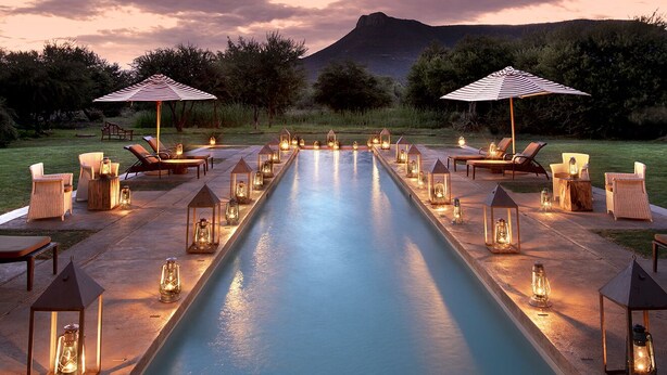 Une piscine entourée de chaises longues et de parasols, illuminée par des lanternes, avec une chaîne de montagnes en toile de fond.