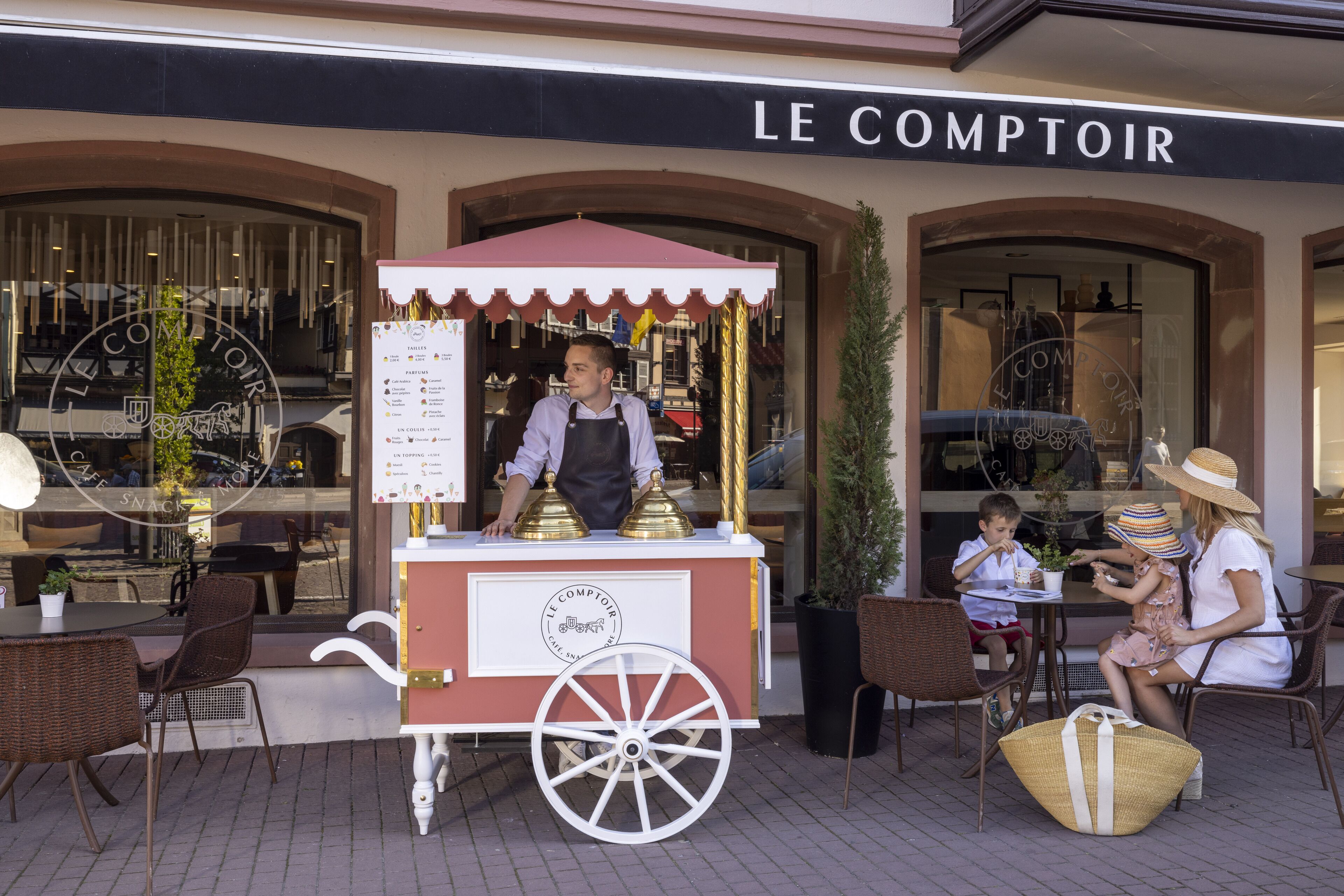 A street food cart outside a cafe named "LE COMPTOIR" with a menu and a person serving.