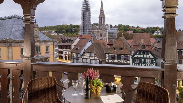 Un balcón con sillas de mimbre, una mesa con copas de vino, una cámara y un jarrón de flores.