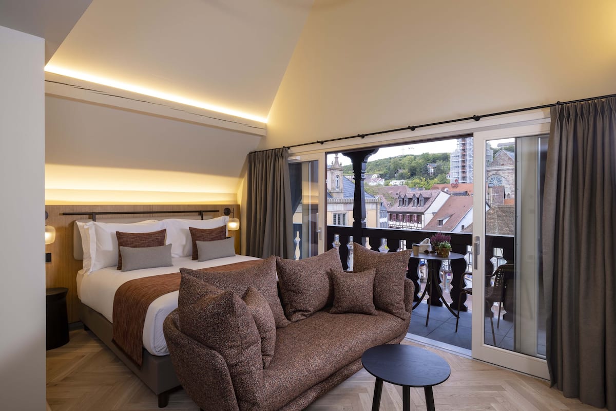 A hotel room with a bed, a brown sofa, a small table, and a balcony with a view of buildings.