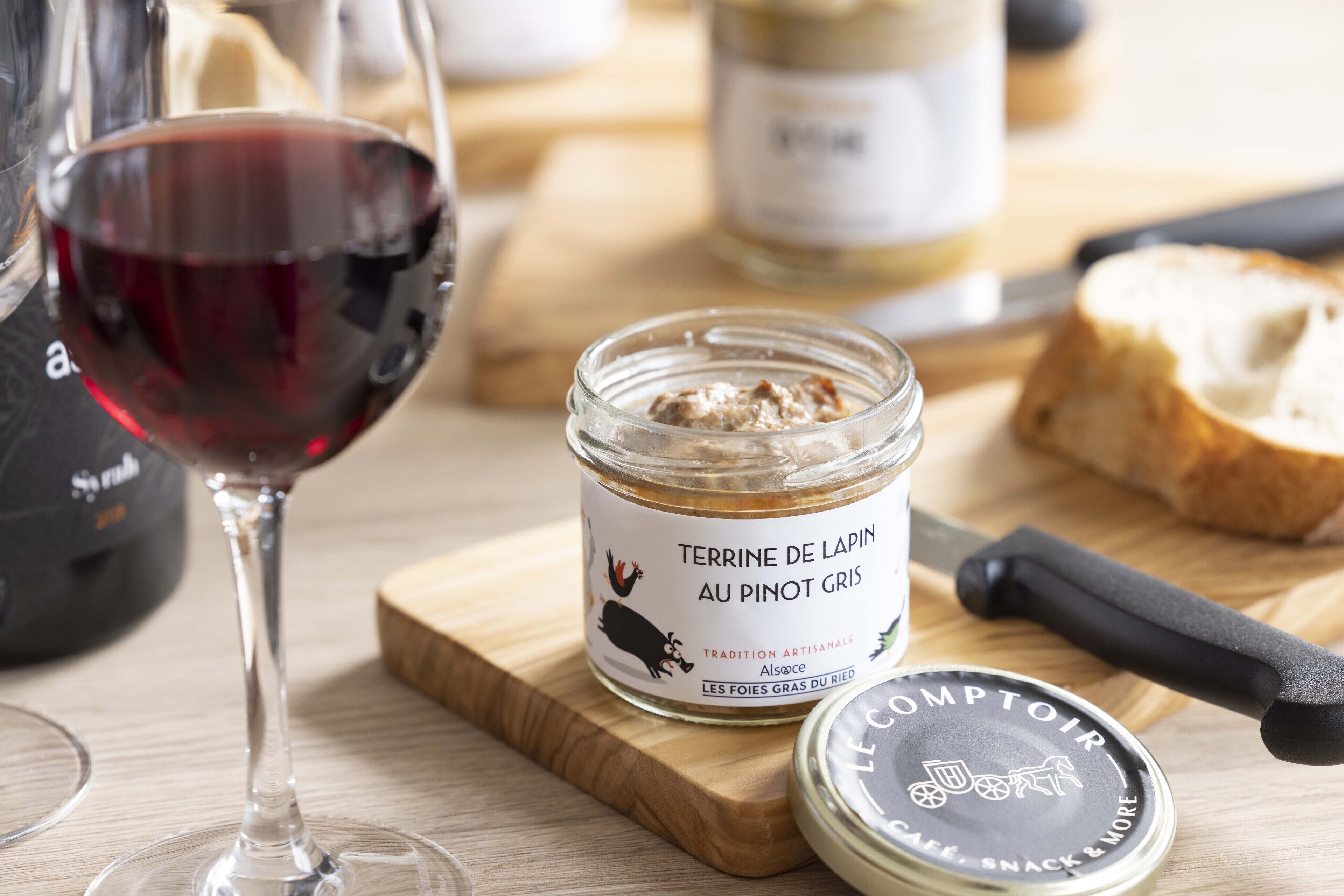 A wooden cutting board with a jar of terrine de lapin au pinot gris, a knife, and a glass of red wine.