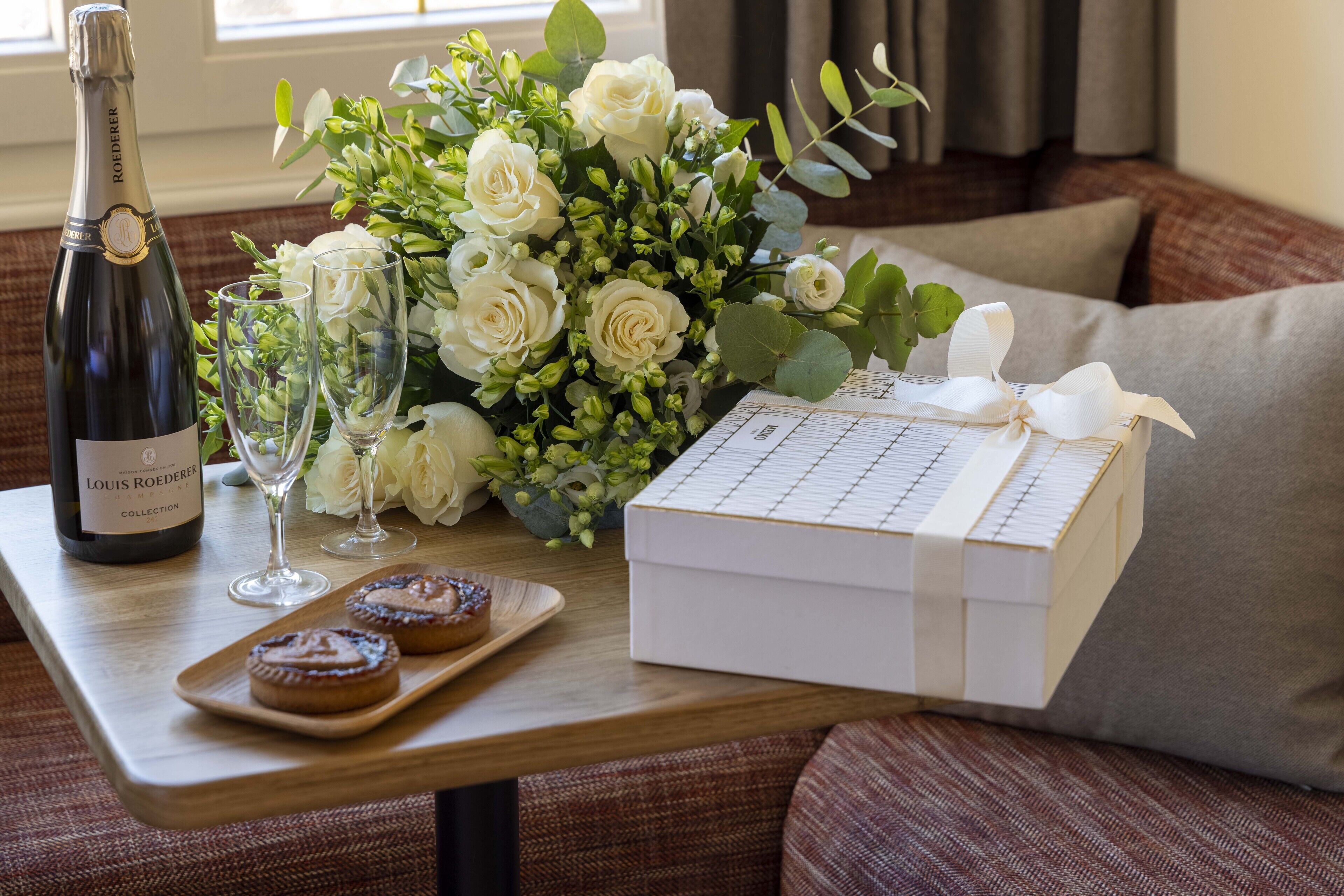 Ein Tisch mit einer Weinflasche, zwei Gläsern, einem weißen Blumenstrauß, einer Schachtel mit Schleife und zwei Pâtisseries auf einem Tablett.