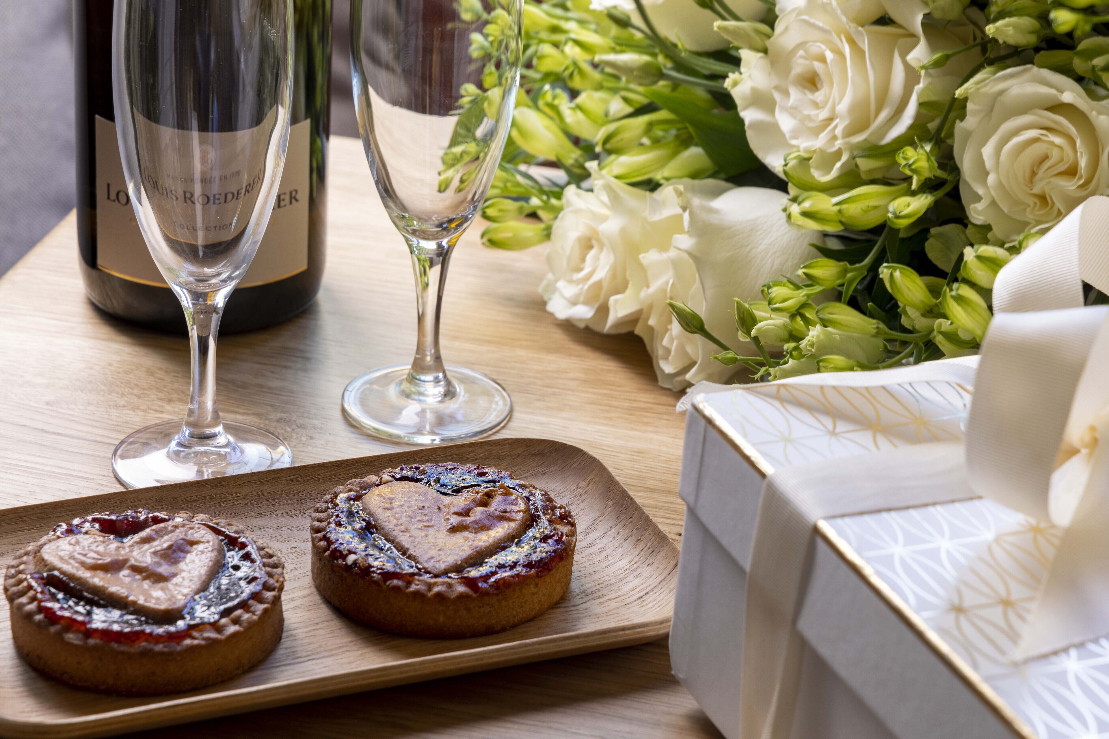 Uma mesa de madeira com dois doces em formato de coração, uma garrafa de vinho, duas taças de vinho e um buquê de flores brancas.