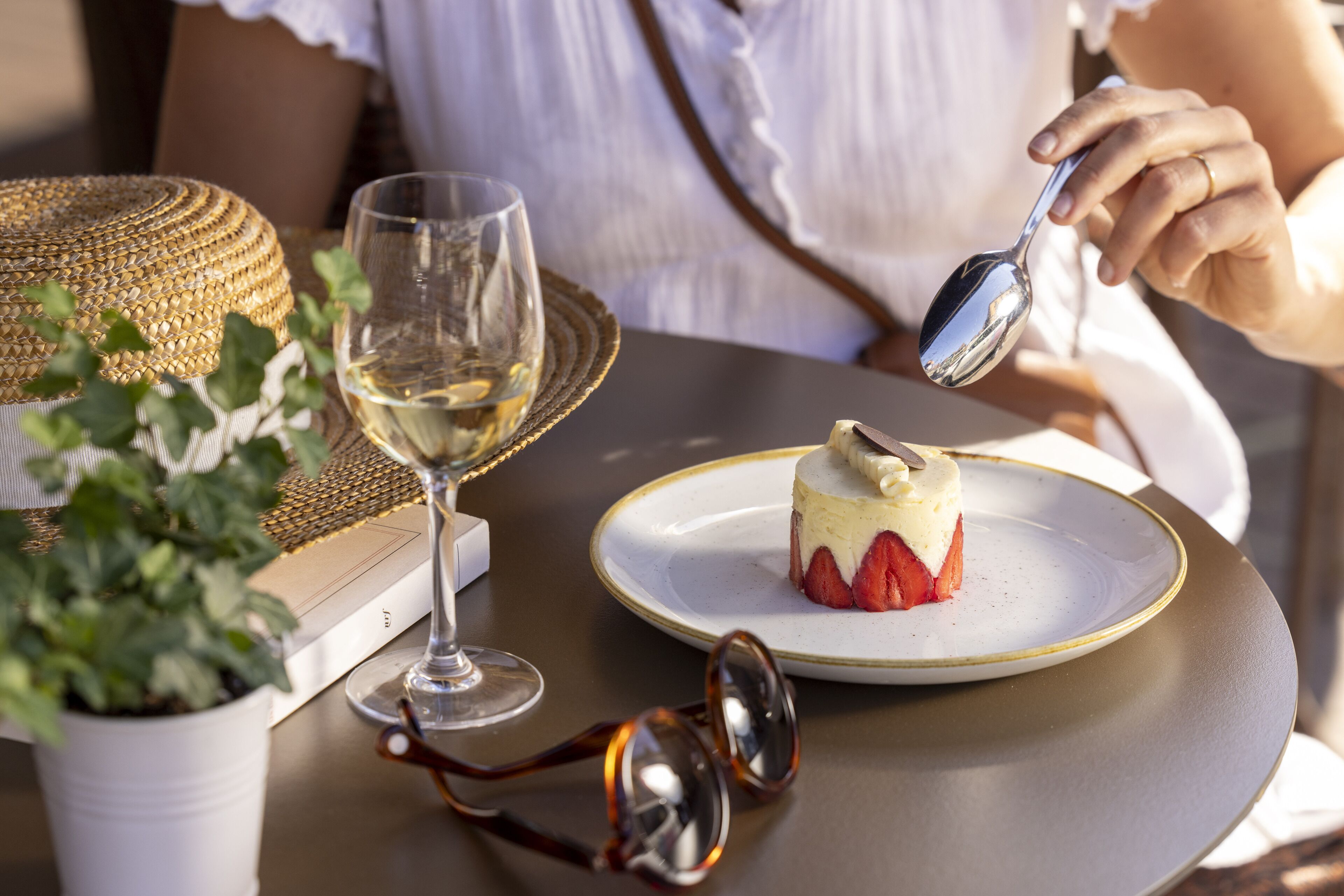 A dessert on a plate with a spoon, a glass of white wine, and a potted plant on a table.