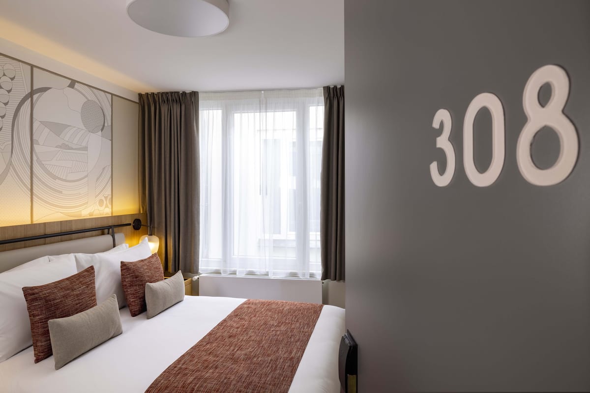 A hotel room with a large bed, a headboard with a decorative panel, a wall-mounted number '308', and a window with curtains.