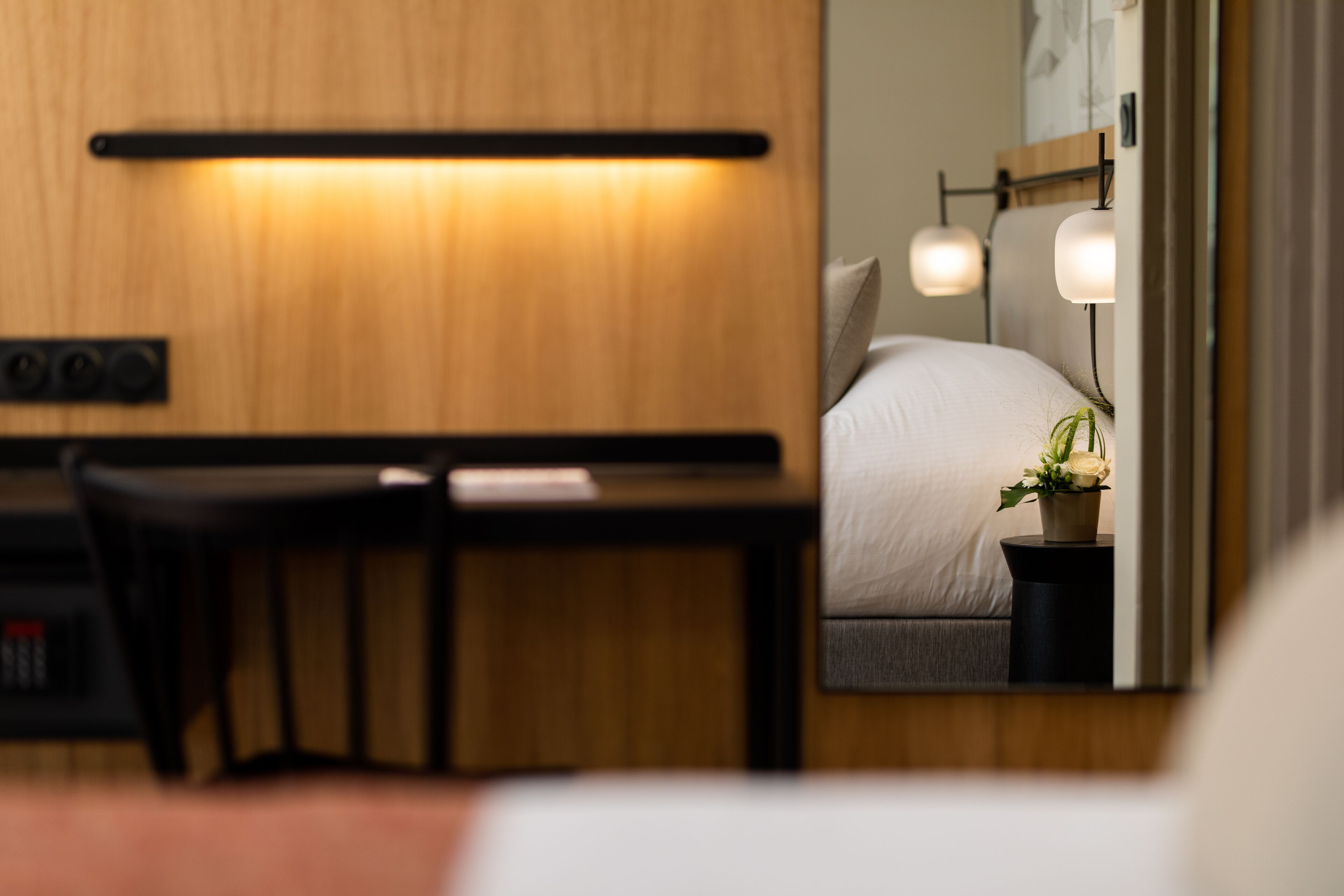A hotel room with a wooden desk, a bed with a white comforter, and a small potted plant on a bedside table.