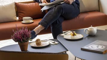 Una persona sentada en un sofá con una taza y un plato, una mesita con una tetera, un plato de comida y un libro.