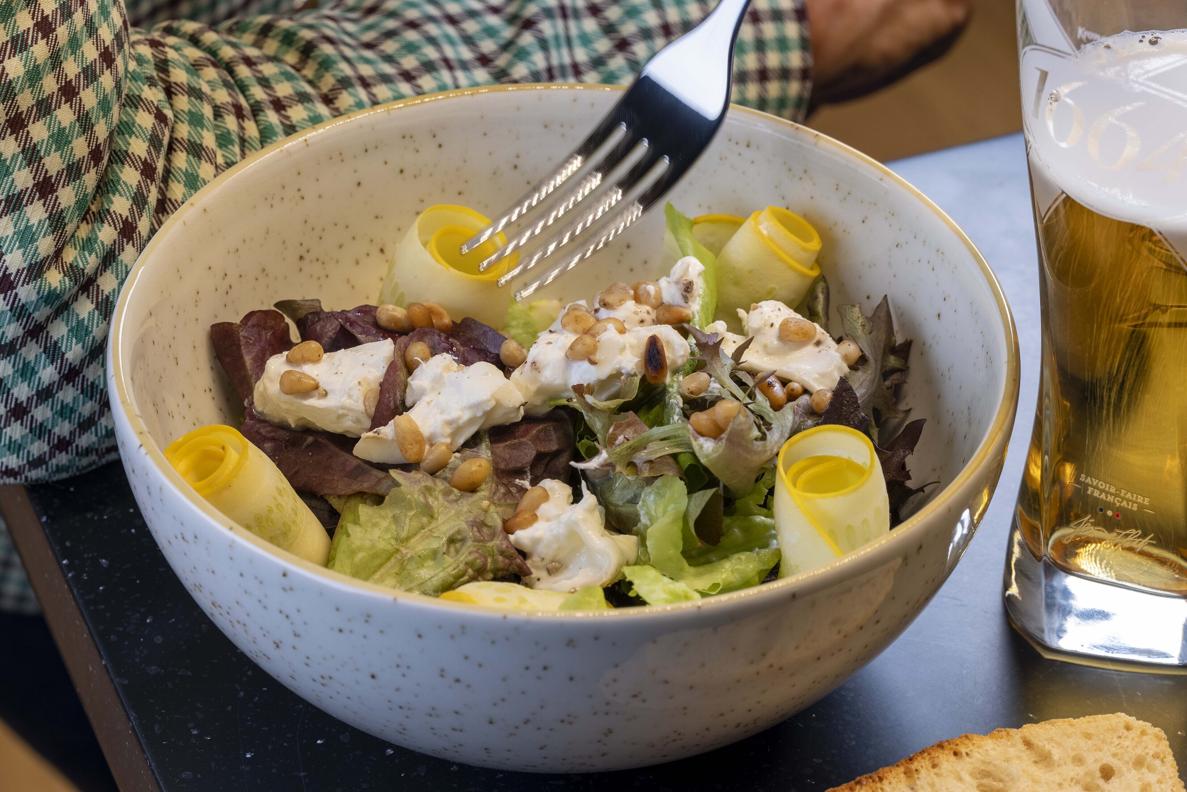 A bowl of salad with a fork, a glass of beer, and a slice of bread on a table.