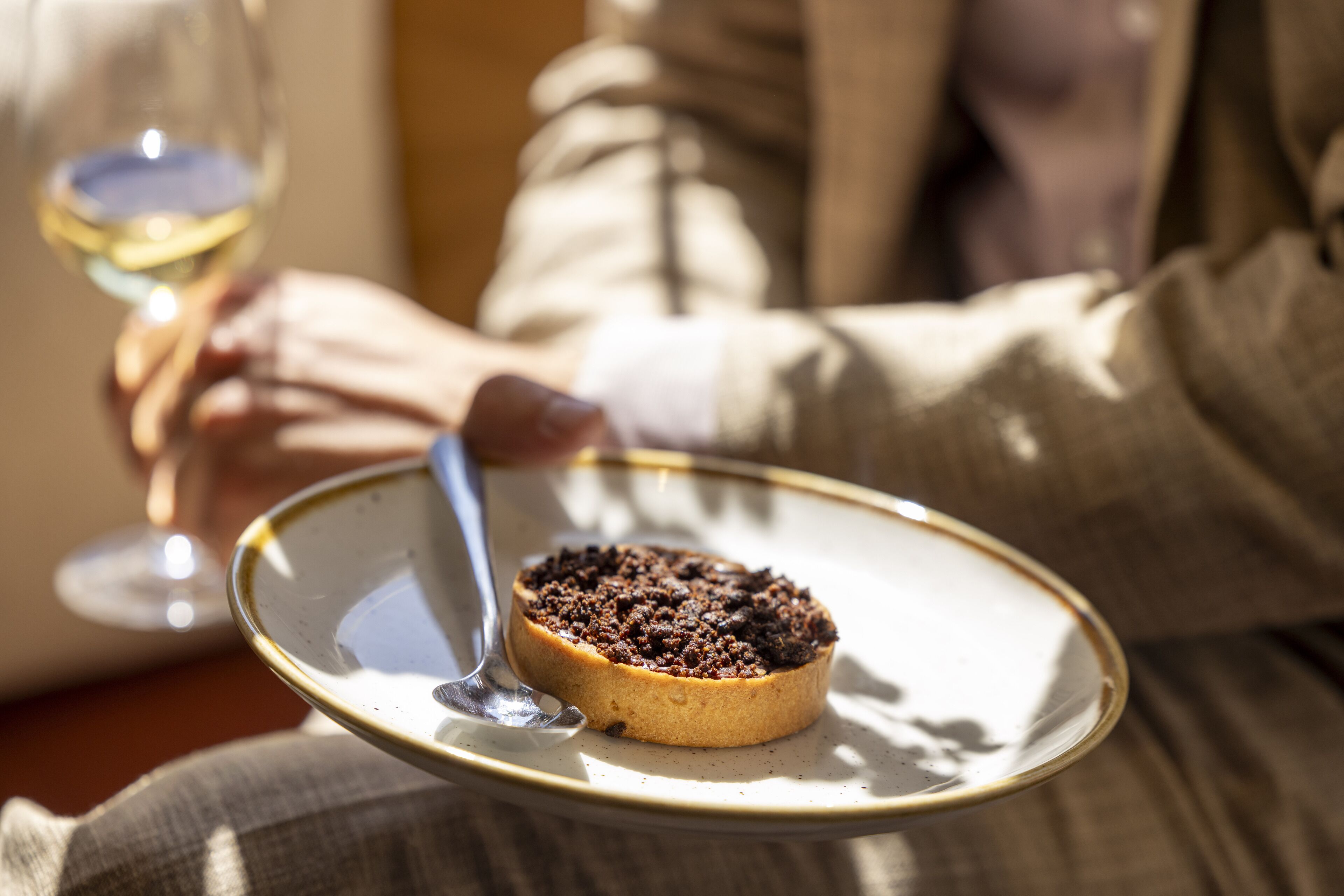 Una persona sosteniendo un plato con un postre y una cuchara, y una copa de vino blanco al fondo.