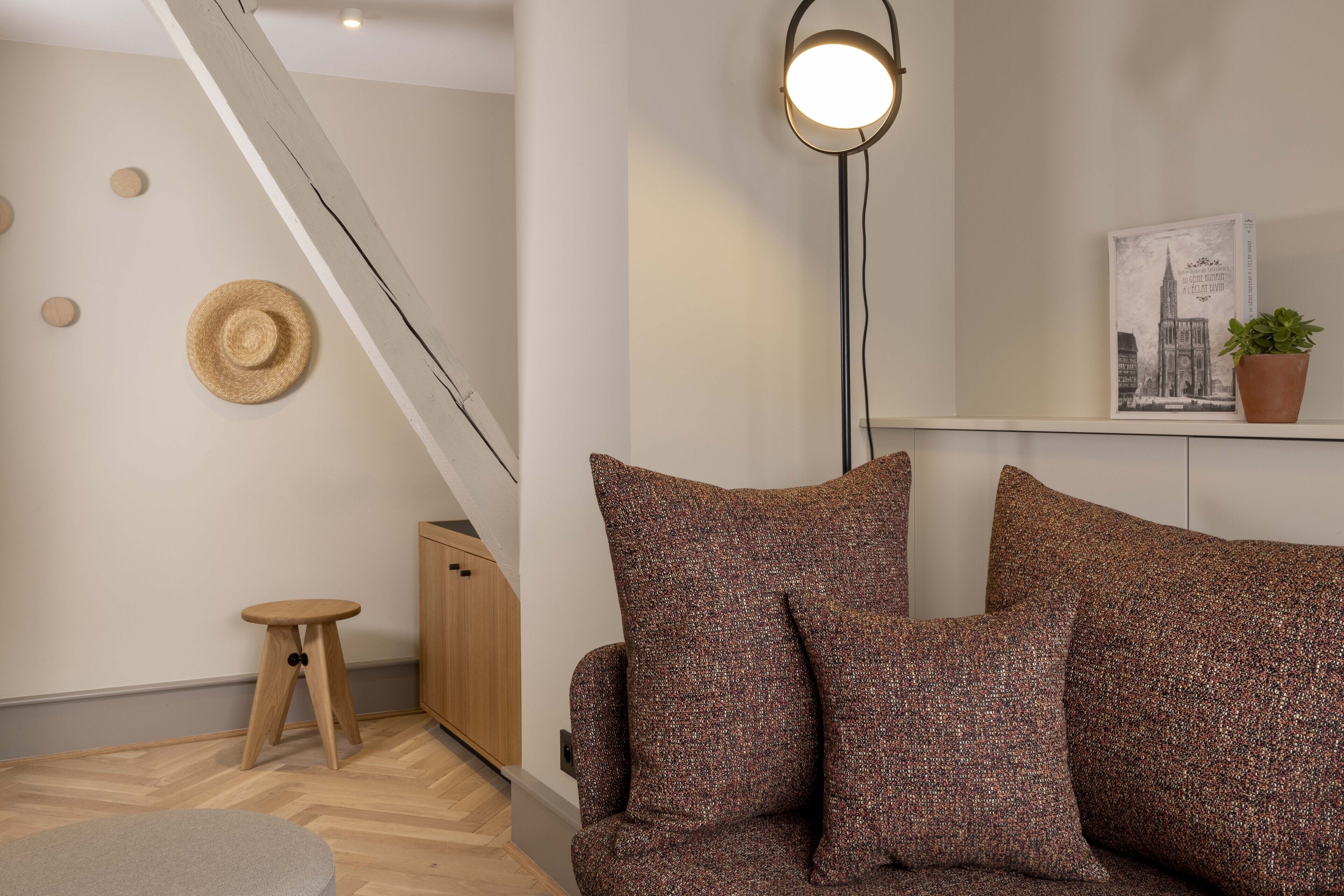 A modern living room with a sofa, a small wooden stool, a hanging hat, a floor lamp, and a framed picture on the wall.