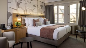 Habitación de hotel con una cama grande, mesitas de noche, una pequeña mesa redonda y vistas al exterior a través de amplios ventanales.
