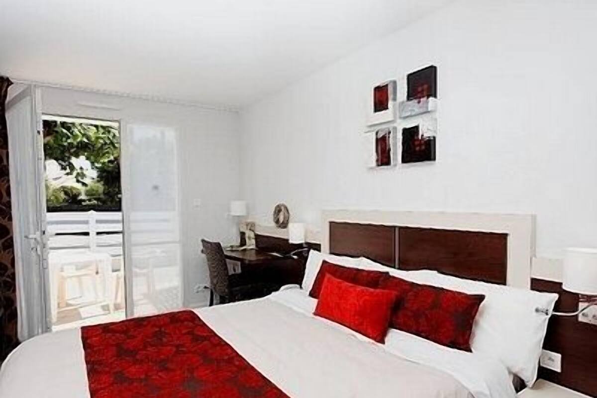 A hotel room with a bed, a desk, and a chair. There is a window with a view of greenery outside.