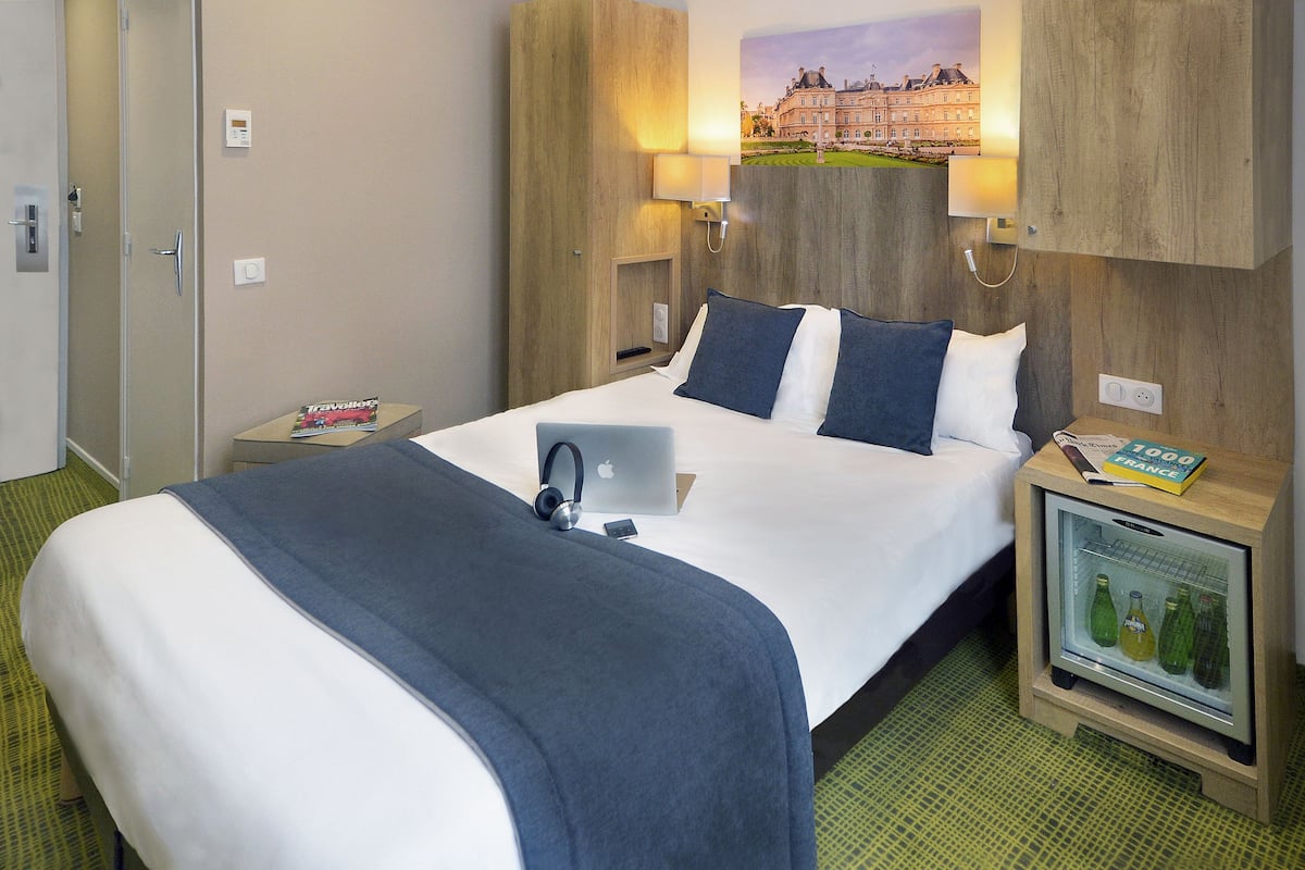 Une chambre d’hôtel avec un lit, un bureau avec un ordinateur portable, un mini-réfrigérateur contenant des bouteilles et une lampe fixée au mur.