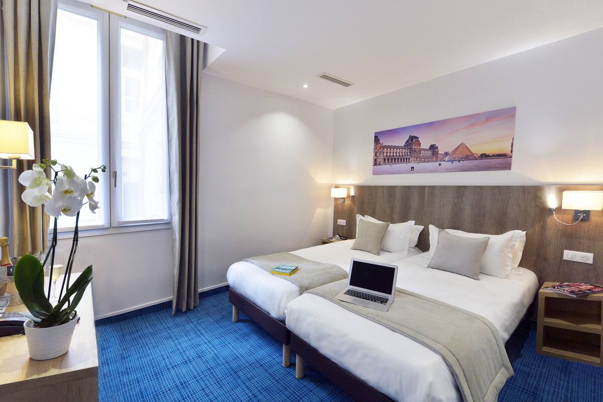 Une chambre d’hôtel avec deux lits, un ordinateur portable posé sur l’un des lits, un tableau représentant un célèbre monument accroché au mur, une plante sur le bureau et une moquette bleue.