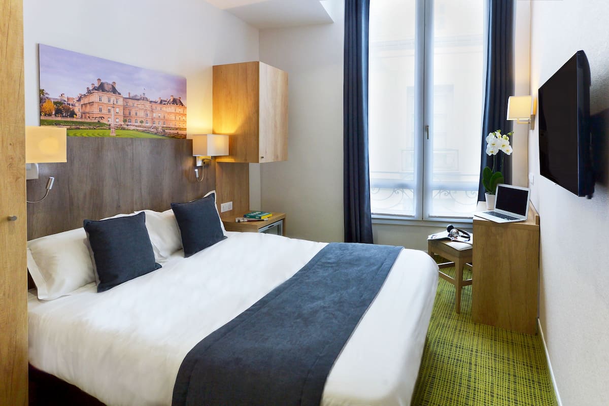 Une chambre d’hôtel avec un grand lit, un bureau avec un ordinateur portable, une télévision et un tableau représentant un bâtiment accroché au mur.
