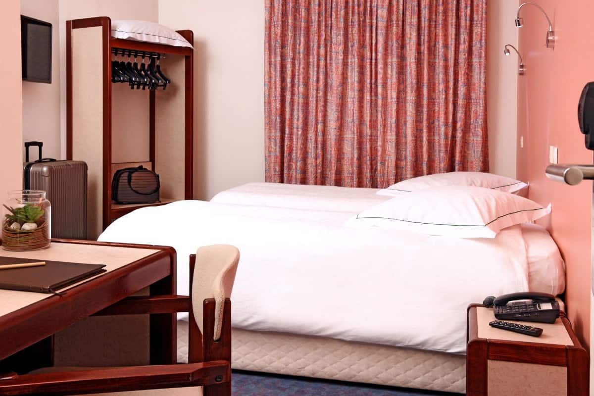 A hotel room with a bed, a desk with a chair, a coat rack, a suitcase, and a remote control on a nightstand.