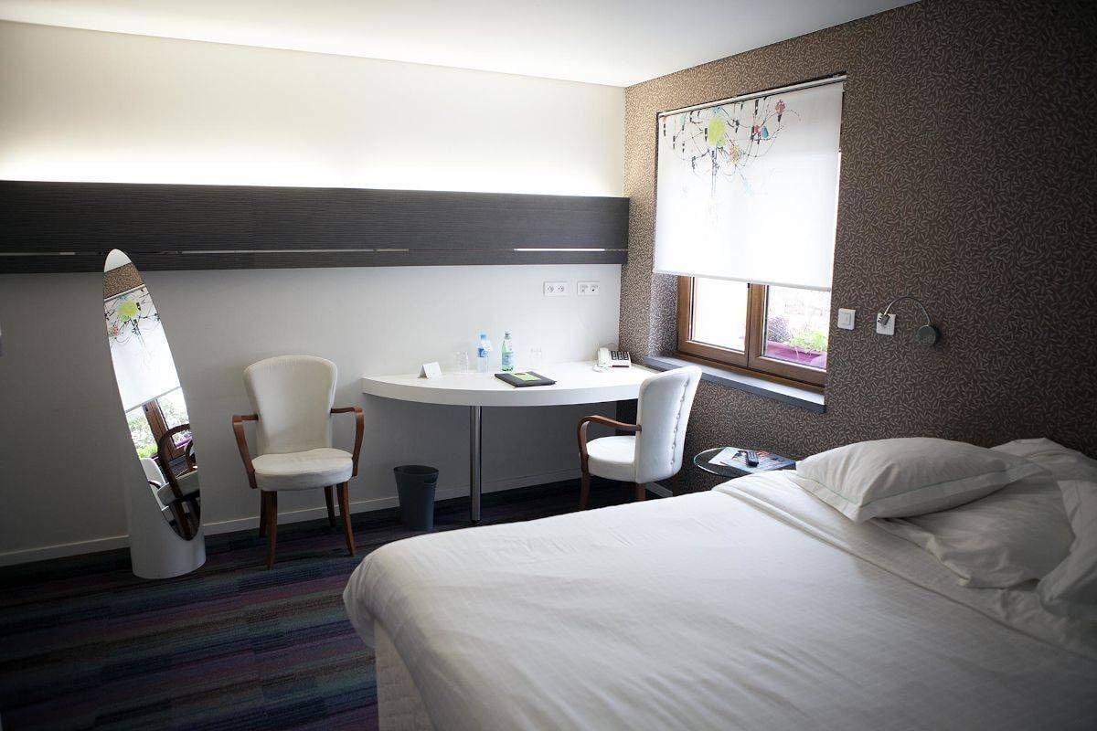 A hotel room with a bed, two chairs, a small table, a window, and a wall with a decorative panel.